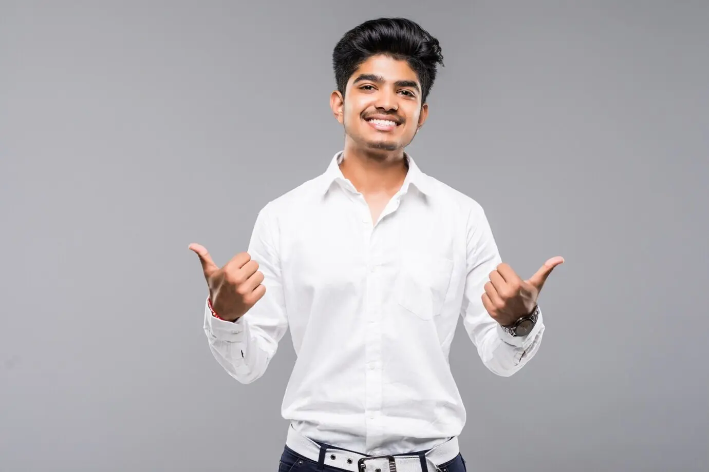 Young Indian man standing against an isolated grey wall, showing approval with a thumbs-up, smiling and happy about his success.