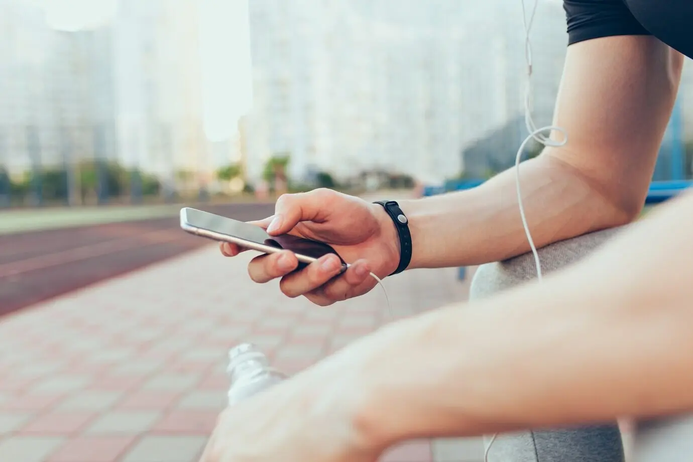 A guy sitting in the city in the morning has a phone in his muscular hand. He is holding a bottle of water and headphones.